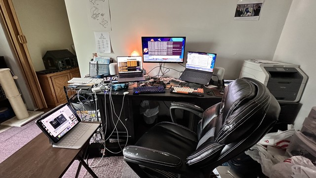 Messy desk with two laptops (14” Apple MacBook Pro, 15” Lenovo Yoga, with a wall-mounted 24-inch LG monitor in the center.)  11-inch Apple iPad Air. on a small table to the left. Brother color laser printer off to the side.  Messy cabling on the left. Various detritus (Raspberry Pis, HomePods, many keyboards) scattered around.