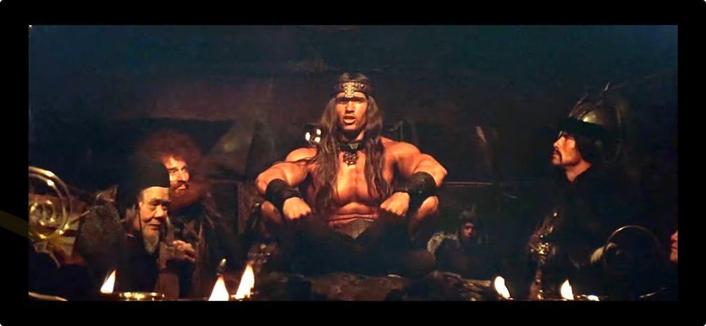 A still image from the movie Conan the Barbarian, showing Conan (Arnold Schwarzenegger) seated, shirtless, and surrounded by other figures in a dimly lit setting. Candles are burning in front of them. Conan is delivering his "What is best in life…?" speech.