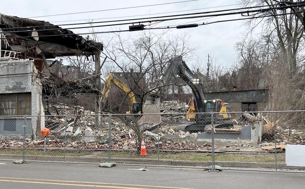 View through a chain-link fence of a two-story building being nibbled to pieces by  heavy equipment