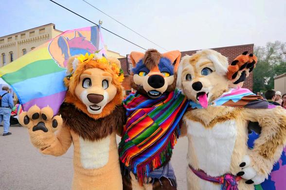 A photo of three fursuiters, a lion, coyote, and dog. The yote has a multi-color kerchief, and the dog wearing a pride flag.  