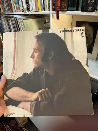 The photo shows someone holding a vinyl record album titled “Stephen Stills 2” by Stephen Stills. The cover features a profile image of Stephen Stills sitting at a table, looking out of a window with his arms resting on the tabletop. He has medium-length hair and sideburns, wearing a dark, relaxed-fit garment. The photo is warm-toned and has a contemplative mood.

In the background, you can see a bookshelf filled with books, LP records, and some framed art or photographs — including a recognizable image of Albert Einstein. The setting gives off a cozy, vintage, and intellectual atmosphere. [lololol]