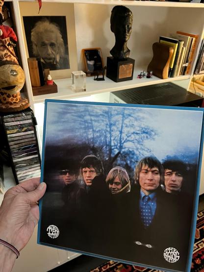 This photo shows a hand holding a vinyl record album by The Rolling Stones. The album cover features a blurred, dreamlike image of the five band members standing outdoors in front of leafless trees under a moody, blue-tinted sky. The photo is stylistically distorted, with the left side especially fuzzy, contributing to a surreal or psychedelic effect.

In the bottom corners of the cover are the logos for “ABKCO Records” and “The Rolling Stones.” This is the 1967 album “Between the Buttons.” [It's a late-model re-release.]

In the background, there’s a well-curated shelf filled with eclectic items:
	•	A framed black-and-white photo of Albert Einstein
	•	A bronze bust sculpture
	•	Small framed photos and objects
	•	A carved wooden head wearing a hat on the left
	•	A stack of compact discs
	•	Books and binders

The space appears cozy, intellectual, and full of character — reflecting both artistic taste and a deep appreciation for music and culture. [ha ha ha yay me]