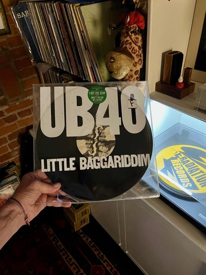 This photo shows a hand holding a picture disc vinyl record of UB40’s album “Little Baggariddim.” The disc is packaged in a clear plastic sleeve, and the artwork on the vinyl itself features bold white text on a black background. The album title, LITTLE BAGGARIDDIM, is printed in all caps below the large “UB40” band logo. In the middle of the vinyl is a small black-and-white photo of the band.

A green sticker on the front promotes hit songs from the album, reading:
“Includes the hit singles: I GOT YOU BABE / DON’T BREAK MY HEART.”

In the background:
	•	You can see shelves filled with vinyl records.
	•	A quirky, painted papier-mâché or carved wooden head figure stands on a shelf nearby.
	•	There’s a “Radiation Records” slipmat on a turntable platter to the right.
	•	The space has a warm, eclectic, and music-focused vibe, with exposed brick, artistic decor, and a cozy, collector’s ambiance.