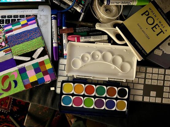 This photo captures a creative workspace, likely an artist's [oh ha ha you flatter me!] desk, filled with various art supplies and personal items.

In the foreground and center, there's an open watercolor paint set, featuring two rows of vibrant, solid watercolor pans. Above it rests a white plastic mixing palette, which appears to be empty and clean, ready for use.

To the left, there are two colorful, hand-decorated or painted panels, possibly made of cardboard or thick paper, showcasing geometric designs and abstract patterns. One has a grid-like pattern of squares in various colors, while the other features stripes and more organic shapes.

Scattered around these central items are several other tools and objects. A pair of white-handled scissors and a roll of white tape are visible near the mixing palette. Various markers and pens, including Sharpies in different colors (purple, blue, red, black, green), are also present. Some are lying on what appears to be a laptop keyboard, which is partially visible on the left side of the frame.

On the right side of the image, part of a black computer keyboard is visible, and on top of it, a book titled "WHEN I WAS A POET" by David Meltzer, from "THE POCKET POETS" series, is open.

The overall impression is one of an active and engaged creative process, with a mix of tools for painting, drawing, and possibly writing or other artistic endeavors.