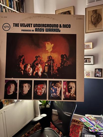 
This is a close-up photo of the front cover of The Velvet Underground & Nico album, produced by Andy Warhol. The album cover is held up, obscuring most of the background, but hinting at a room with shelves and artwork.

The album cover itself is prominently displayed. At the top left, the "Verve" record label logo is visible. The title "THE VELVET UNDERGROUND & NICO" is printed in large black letters, with "PRODUCED BY ANDY WARHOL" directly below it. "STEREO" is also printed in the top right corner.

The main image on the cover is a dark, somewhat abstract group portrait of the band members, bathed in reddish and orange hues. Lou Reed is centrally positioned, looking up.

Below the main band photo, there are five individual, square portraits of the band members, each with a distinct, slightly blurred or painterly quality, typical of the era's photographic techniques and Warhol's influence. From left to right, these appear to be John Cale, Sterling Morrison, Maureen Tucker, Lou Reed, and Nico.

The background, partially visible around the edges of the album, suggests an indoor setting. On the left, there are shelves filled with books. On the right, there's a dark blue wall with several framed artworks hanging on it. The bottom of the image shows a hint of a chair and a colorful rug or mat on the floor.