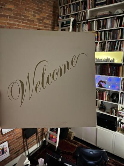 This photo features a piece of light-colored cardstock or paper with the word "Welcome" elegantly written in a script font across its center. The "Welcome" sign is being held up, largely filling the foreground of the image. [It's an album cover, Santana is the lead artist.]

The background reveals an indoor setting, likely a room with a warm, inviting atmosphere. The dominant feature in the background is a large, tall white bookshelf filled with numerous books, stretching almost to the ceiling. The shelves appear well-stocked, suggesting a personal library or study.

To the left of the bookshelf, a section of an exposed brick wall is visible, adding a rustic or industrial touch to the space. Below the bookshelf, there are white cabinets and some electronic equipment, including what looks like a monitor or speaker.

On one of the lower shelves of the bookshelf, there's a small, illuminated display of items, possibly decorative figures or other objects, creating a cozy glow. The floor below shows a patterned rug in warm tones, contributing to the lived-in feel of the room.

Overall, the photo conveys a sense of hospitality and a personal space filled with books and character.