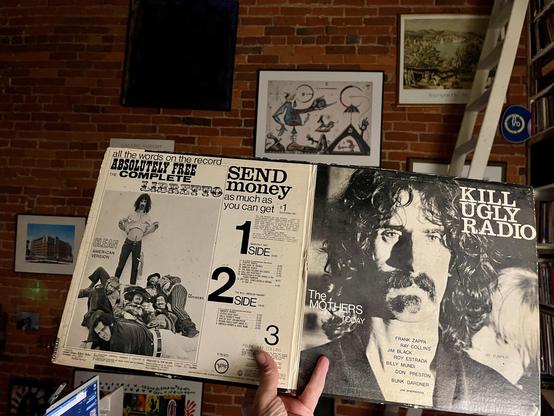 This photo shows the open gatefold of the The Mothers of Invention - Absolutely Free vinyl album, revealing its internal artwork and text. On the right panel of the gatefold, the album cover's original artwork is visible: a large, striking black and white portrait of Frank Zappa with his distinctive mustache and beard, looking directly at the viewer. Below his image, the text "THE MOTHERS TODAY" and a list of band members are present. The phrase "KILL UGLY RADIO" is prominently displayed in a bold, stylized font towards the top right of this panel.

The left panel of the gatefold contains various textual and graphical elements, designed to look like a "LIBRETTO" or manifesto. It features the title "ABSOLUTELY FREE" and mentions "all the words on the record." There are sections with a "CLEAN LYRIC VERSION" and a "SEND money as much as you can get +$1" offer.     The tracklisthoto provides a glimpse into a room with an artistic and well-read aesthetic. A brick wall dominates the left side, adorned with several framed artworks. One dark, abstract piece is prominently centered on the brick. Further to the right, a large white bookshelf filled with books stretches vertically, and a white ladder is leaning against it, suggesting it's used to reach higher shelves. The overall impression is of a rich, lived-in space that blends art, music, and literature. [ooh la la]