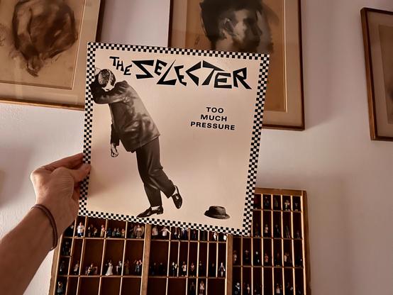 This photo displays the album cover for The Selecter - Too Much Pressure, held by a hand on the left. The cover features a black and white checkerboard border, framing a stylized black and white image of a person in a suit and hat, leaning forward as if against a strong force. The band's name, "THE SELECTER," is in a distinctive font at the top, and "TOO MUCH PRESSURE" is centered on the right.

The background shows a wall with several framed artworks, primarily portraits or busts, in warm, neutral tones. Below, a wooden display cabinet with many small compartments, each holding miniature figures or objects, adds to the room's eclectic decor.