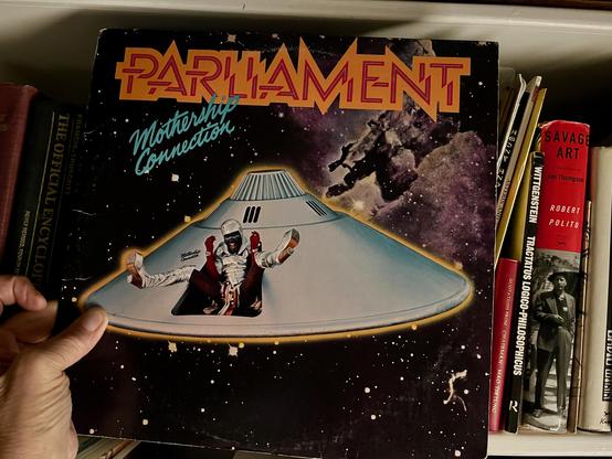 This photo features the album cover for Parliament - Mothership Connection, held by a hand on the bottom left. The album is tilted slightly, showcasing its vibrant and iconic artwork.

The cover depicts a large, light grey, saucer-shaped spaceship with a smaller, cone-like structure on top, resembling a UFO. Emerging from an opening in the spaceship is a figure dressed in a white and silver, futuristic-looking costume, with arms outstretched, seemingly "landing" or "emerging" from the vessel. The background is a dark, starry cosmic scene with nebulae or distant galaxies.

Above the spaceship, the word "PARLIAMENT" is written in large, stylized, orange and yellow block letters. Below it, in a cursive, lighter font, is "Mothership Connection."

The album is nestled among other books and records on a shelf, which forms the background of the image. Visible book spines include "SAVAGE ART ROBERT POLITO," "WITTGENSTEIN TRACTATUS LOGICO PHILOSOPHICUS," and "Quotations from Chairman Mao." This suggests the album is part of a larger collection within a library or personal study space.