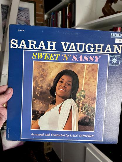 This photo prominently features the album cover for Sarah Vaughan - Sweet 'n' Sassy, held by a hand on the bottom left. The album cover is predominantly dark blue.

In the center of the cover, there is a large, rectangular framed portrait of Sarah Vaughan. She is smiling gently at the viewer, wearing a light-colored, possibly white or cream, draped top that exposes one shoulder. Her hair is styled in a classic, elegant bob. The background of her portrait is an abstract blend of warm, earthy tones, suggesting an outdoor or natural setting.

Above her portrait, in large white letters, is "SARAH VAUGHAN". Below her image, in yellow text, is the album title "SWEET 'N' SASSY". At the very bottom of the framed portrait, "Arranged and Conducted by LALO SCHIFRIN" is credited.

In the top right corner of the album cover, "STEREO" and the "ROULETTE BIRDLAND SERIES" logo are visible. A price tag of "$5.00" is also present in the top right. The catalog number "SR 52112" is in the top left corner.

The background of the photo is somewhat out of focus but shows a room with a bookshelf filled with books on the right, and hints of a brick wall and other decor on the left, similar to the setting in previous images. The overall impression is that the album is part of a personal collection in a well-decorated space. [oh you flatterbot, you]