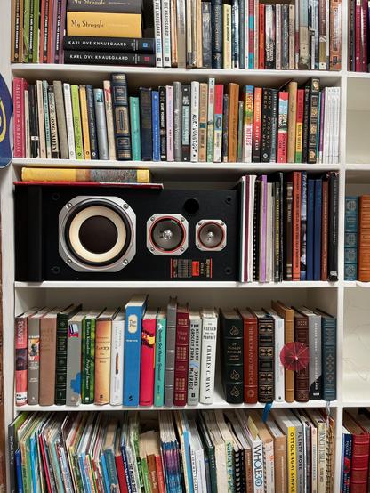 This photo shows a tall, white built-in bookshelf filled with a large and eclectic collection of books, with a vintage Fisher STV-103 speaker prominently occupying one of the central cubbies.

In the center of the photo, a black Fisher speaker with silver and red accents featuring three drivers (woofer, midrange, tweeter) and a ported enclosure is integrated into the bookshelf layout, occupying an entire shelf horizontally. 

Above the speaker a bright yellow Van Gogh art book lies horizontally…