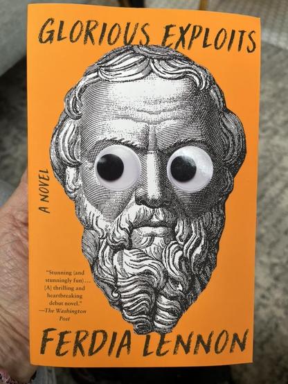 This photo shows the cover of the novel Glorious Exploits by Ferdia Lennon. The book has a bold, bright orange background, and features a black-and-white illustration of a classical bust or statue—most likely depicting a Greek philosopher or playwright, in keeping with the book’s theme.

What makes the cover humorous and eye-catching is the addition of large googly eyes stuck onto the statue’s face, giving the ancient figure a whimsical, almost absurd expression.

Text on the cover includes:

•	Title at the top: “GLORIOUS EXPLOITS” in a bold, handwritten style.
•	Subtitle: “A NOVEL” along the left side.
•	Author’s name: FERDIA LENNON at the bottom in matching handwritten font.
•	A blurb from The Washington Post on the left:“Stunning (and stunningly fun)… [A] thrilling and heartbreaking debut novel.”

The hand holding the book suggests the image was taken in a bookstore or library, possibly over a carpeted or tiled floor. The googly eyes add a layer of irreverent fun to what is likely a literary or historical novel.