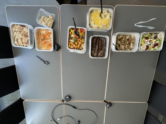 An overhead view of a variety of Mediterranean or Middle Eastern-style dishes arranged in aluminum trays on a large conference table. The food appears to include:

	•	Pita bread slices
	•	A creamy orange curry or stew (possibly butter chicken or chicken tikka masala)
	•	A tray of baklava (dessert)
	•	Grilled vegetables with skewered chicken or paneer
	•	Shredded rice pilaf or biryani with saffron and dried fruit
	•	Grilled koobideh or kebab meat
	•	A tray of a saucy spinach and rice dish (possi…