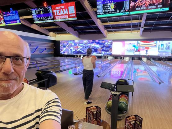 This photo was taken inside a bowling alley. In the foreground on the left, a man is taking a selfie—he has glasses, a white shirt with dark blue horizontal stripes, and a small goatee. Behind him, a woman is seen from the back holding a bowling ball, preparing to bowl. She’s wearing a sleeveless white top and dark pants.

There are multiple bowling lanes stretching toward a large wall-sized screen at the far end, which is displaying a split-screen TV broadcast (one side appears to show a footb…