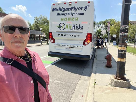 This photo shows a man taking a selfie near a Michigan Flyer bus. He is wearing a red button-down shirt over a black t-shirt, sunglasses, a beaded necklace, and a black crossbody strap (likely for a bag). Behind him, the Michigan Flyer bus is stopped at a curb, with several people boarding or unloading near the front. The bus prominently features the AirRide logo and indicates its route includes Lansing, Brighton, Ann Arbor, and Detroit Metropolitan Airport (DTW).

Street signage is visible, al…