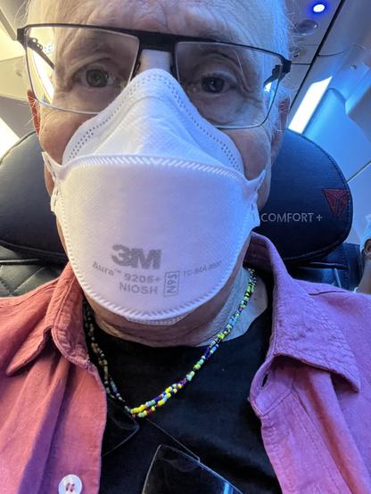 This is a close-up selfie of a man seated on an airplane. He is wearing a white 3M Aura 9205+ NIOSH N95 respirator mask, black-rimmed glasses, a multicolored beaded necklace, a black shirt, and a red button-down shirt layered over it. The background shows the airline seat’s headrest, which has the Delta Comfort+ branding, confirming he is flying with Delta Air Lines in the Comfort+ section. Overhead lights and part of the cabin ceiling are also visible, with a blue-tinted lighting typical of airplane interiors during boarding or cruising.