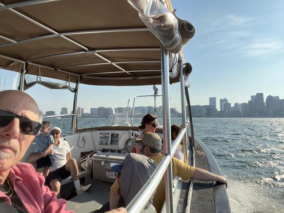 The image depicts a group of people enjoying a boat ride on the water, with the skyline of Boston visible in the background under a clear sky. Several individuals are seated or standing on the boat, including an older man in the foreground wearing sunglasses [it me!] and a younger child being held by an adult. The boat is moving, creating a wake in the water, and the sun appears to be shining brightly. The Boston skyline, featuring numerous buildings, is clearly visible across the water.