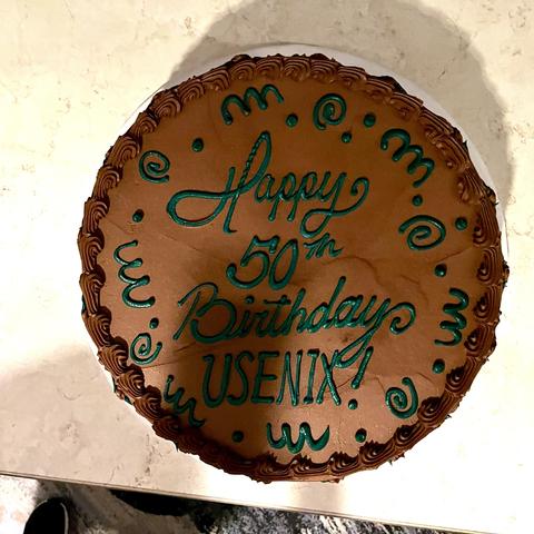 This is a photo of a circular birthday cake with chocolate frosting. The cake has a decorative border of piped chocolate rosettes or swirls around the edge. The main surface is covered in smooth chocolate frosting with text written in teal or green piping that reads “Happy 50th Birthday USENIX!” The text is surrounded by decorative flourishes and dots in the same teal color. The cake appears to be sitting on a white surface, and you can see part of what looks like a person’s dark clothing at the bottom of the image. This appears to be a celebration cake for USENIX’s 50th anniversary.​​​​​​​​​​​​​​​​