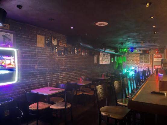 This photo shows the interior of a cozy, dimly lit bar or jazz/blues club with a vintage and intimate atmosphere. Key elements include:
	•	Exposed brick walls with framed portraits and memorabilia, likely of musicians or historic patrons.
	•	A neon-lit jukebox or digital music station on the left wall, glowing with purples and pinks.
	•	Rows of wooden tables and chairs, arranged closely together for a communal vibe.
	•	A long wooden bar with stools along the right side of the image.
	•	A small stage setup at the far end, equipped with a drum kit illuminated by green, blue, and red stage lights.
	•	Signs on the back wall reading “EMERGENCY EXIT ONLY” add a functional touch.
	•	The lighting is low overall, but accentuated by moody neon, giving it an atmospheric and inviting feel—perfect for live music.

This venue looks like a classic, no-frills spot where live music—possibly jazz, blues, or rock—is performed regularly.