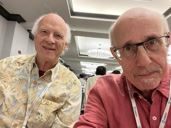 This photo shows Rio Farrow on the left and me on the right, seated indoors at the conference. We are bothwearing lanyards with “USENIX” printed on them — we are attending a USENIX conference (a well-known association focusing on advanced computing systems).
	•	Rik is smiling warmly and is wearing a patterned shirt with light earthy tones.
	•	I am taking the ussie wearing a red button-up shirt and glasses and a neutral expression.

In the background, you can see a modern conference room with white ceilings, hanging light fixtures, and several other attendees facing away from the camera. The mood is collegial and relaxed.