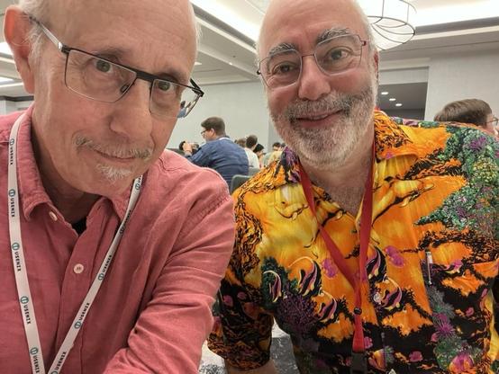 This photo showsme and Dan Klein posing for a cheerful ussie at the conference.
	•	I am again wearing a red button-up shirt and glasses. I have a mustache and goatee, and a white “USENIX” lanyard around my neck.
	•	Dan is smiling broadly and wearing a vibrant, colorful shirt featuring an abstract or tropical landscape with bright oranges, purples, greens, and black. He also has glasses, a graying beard, and a red lanyard.

In the background, other attendees are seated and engaged in conversations at round tables, at a social event during the conference. The room is the same venue as in the previous image, with modern lighting and a professional yet relaxed atmosphere.