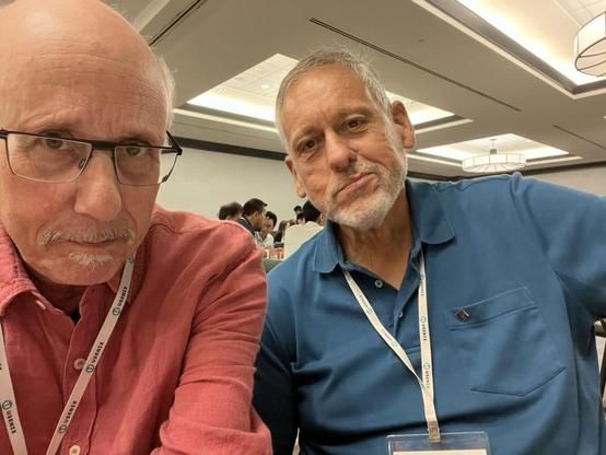 This photo features me and Tom Lyon posing at the conference.
	•	I am still wearing a red button-up shirt, glasses, and a white “USENIX” lanyard. In the photo, I have has a closely trimmed white mustache and goatee and a mildly serious expression.
	•	Pugs is wearing a teal blue polo shirt and also has a “USENIX” lanyard around his neck. He has short gray hair, a trimmed beard, and is smiling slightly.

The setting remains consistent with the earlier photos: a well-lit conference room with modern ceiling lights and several attendees in the background, suggesting a lively and collegial gathering.
