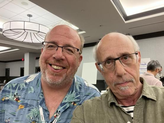 This photo shows me and Brian Noble taking an ussie together in a modern conference space. Brian is wearing a colorful Hawaiian-style shirt with a blue background featuring palm trees and Mickey Mouse. He has gray hair, a full gray beard, black-framed glasses, and is smiling broadly at the camera. I am wearing an olive-colored button-up shirt and have glasses, a gray goatee, and a more subdued expression.

The setting is a contemporary professional space with white walls, a dropped ceiling with fluorescent lighting, and a notable large circular pendant light fixture with an organic, leaf-like pattern. Natural light comes through skylights in the ceiling. [Not quite, Claude.] In the background, you can see other people and a modern meeting area. Both of us are wearing conference badges.
