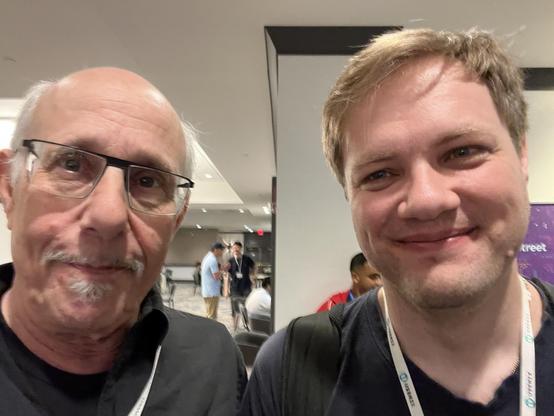 This photo shows me and Jef Sipek taking an ussie in a modern conference or event space. I am older with a bald head, gray goatee, and glasses, wearing a black shirt. Jef is younger with blonde/light brown hair and is wearing a dark gray or charcoal colored shirt. Both of us are wearing conference lanyards with badges.

The setting is a contemporary venue with white walls, modern lighting, and clean architectural lines. In the background, you can see other conference attendees mingling, and there's a purple banner or sign visible on the right side of the frame.