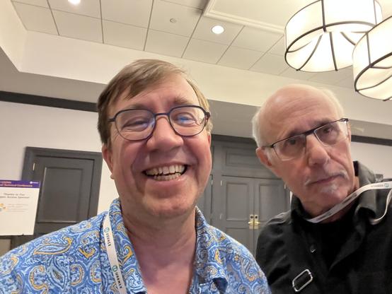 This photo shows me and Keith Smith taking an ussier in a modern conference facility. On the left, Keith is wearing a vibrant blue paisley-patterned shirt and has glasses, with a big genuine smile on his face. He's wearing a conference lanyard. On the right, I am older, wearing a dark sweater or cardigan over a collared shirt, and also have glasses and a more reserved expression.

The setting is a contemporary professional space with white walls, a dropped ceiling with recessed lighting, and distinctive modern pendant light fixtures with an organic, leaf-like design. In the background, you can see gray doors and informational signage.