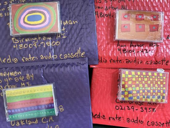 This image showcases several padded envelopes intended for postal shipment, each containing an audio cassette. The envelopes are various colors (purple and red) and have handwritten addresses and "Media rate: audio cassette" inscribed on them, indicating they are being shipped at a discounted postal rate specifically for media. 

Notably, each cassette tape visible in its clear case features a unique and colorful hand-drawn label design, adding a personalized touch to the mailed items. The addr…