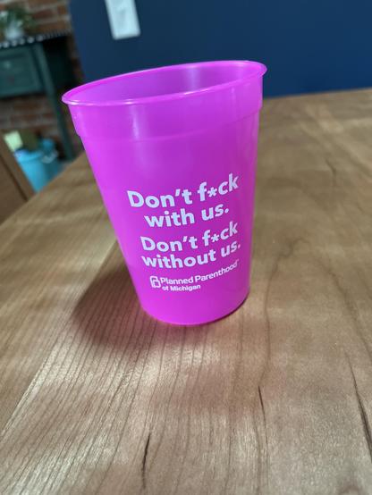 Swag from Planned Parenthood, popular with the grandchildren bc it changes color and none of them can read yet lolol

This photo features a bright pink plastic cup resting on a wooden table. The cup is prominently displayed in the center of the frame and features bold white text with a strong message: "Don't f*ck with us. Don't f*ck without us." Below this, a smaller line of text identifies the organization as "Planned Parenthood of Michigan."