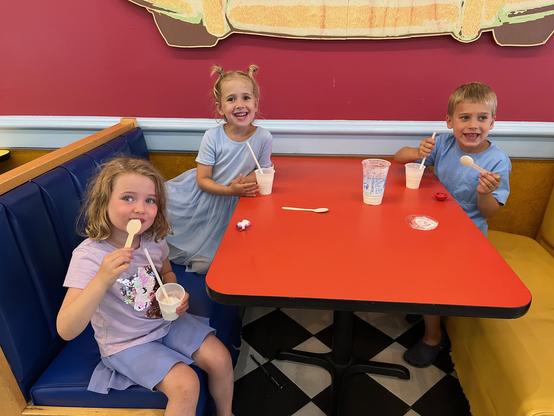A four-, five-, and a six-year old are seated in a booth with a bright orange tabletop in front of a purple each with a small cup containing vanilla milkshake. They're eating with straws and spoons. So far, it is not a messy disaster.

This is a cheerful photo of three children enjoying milkshakes at a restaurant booth. The setting is a classic ice cream parlor with its distinctive blue and yellow booth seating and black-and-white checkered floor.

The children are seated around a bright orange…
