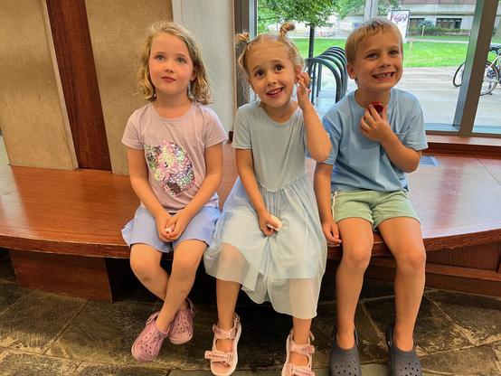 This photo shows three children sitting together on a wooden bench in a public building. The setting has large windows in the background showing green grass and trees outside, with some chairs and bicycles visible through the glass.

On the left is a girl with shoulder-length blonde wavy hair wearing a light pink/lavender t-shirt with a sparkly design paired with light blue shorts and pink Crocs-style shoes. In the middle is a girl with her hair in small buns or pigtails, wearing a light blue d…