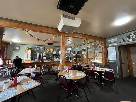 This photo shows the interior of a cozy, small-town restaurant. The atmosphere is warm and homey, with wood-paneled pillars and trim, dark carpeted floors, and simple furnishings.

Several tables with maroon chairs are arranged throughout the dining area. Each table has laminated menus, ketchup and mustard bottles, salt and pepper shakers, and sugar dispensers. The restaurant is decorated with colorful artificial flower garlands strung across the ceiling beams, adding a festive and cheerful tou…