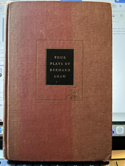 This photo shows the cover of a vintage hardcover book titled “Four Plays by Bernard Shaw.” The cover is a textured reddish-brown cloth with a black rectangular label centered near the top. The title and author’s name are stamped in gold lettering on the label:

FOUR  
PLAYS BY  
BERNARD  
SHAW

A gold-inked rectangular border surrounds the black label, and at the bottom right of the border is a small, stylized design that resembles a publisher’s colophon. The cover shows some wear, indicating …