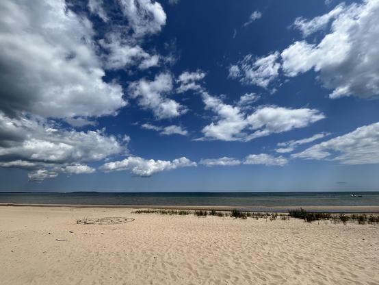 This photo shows a beautiful beach scene with pristine sandy shores stretching toward calm blue-green waters. The sky dominates much of the frame, filled with dramatic white and gray clouds against a deep blue backdrop. The beach appears relatively undeveloped, with natural dune grass growing behind the shoreline and what looks like a simple circular pattern or marking drawn in the sand in the foreground. Small boats or objects can be seen on the distant horizon. The lighting suggests it’s a pa…