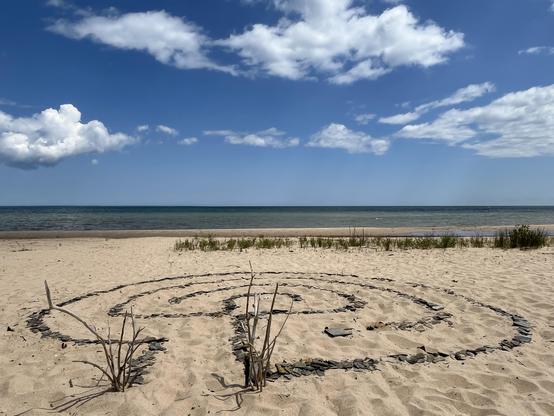 This photo shows the same beach location from a closer perspective, focusing on an intricate spiral pattern in the sand made from dark stones arranged in concentric circular paths forming a walking meditation labyrinth. A few dried plant stems or branches stick up from within the pattern. The beach setting remains consistent with the first image - sandy shores, dune grass in the background, and the same blue-green ocean waters under a partly cloudy sky. This appears to be someone’s artistic cre…