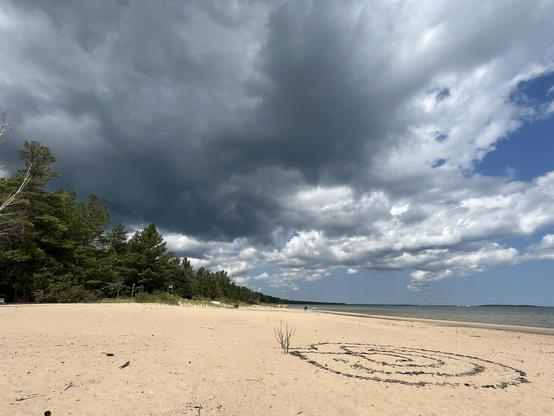 This photo captures the same beach location but with dramatically different weather conditions. The sky is now dominated by dark, ominous storm clouds that create a moody, overcast atmosphere. The lighting is much more subdued and gray compared to the bright, sunny conditions in the previous images. You can see the same spiral pattern in the sand, though it appears less distinct in this lighting. The beach stretches into the distance where there’s a tree line visible on the left side of the fra…