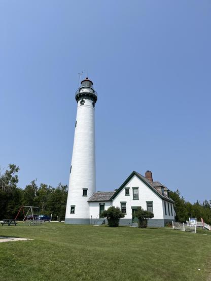 This photo shows a tall, cylindrical white lighthouse with a distinctive lantern room at the top featuring a green/dark colored roof and metal framework. The lighthouse stands prominently against a clear blue sky. Adjacent to it is a white keeper’s house with green trim around the windows and doors, giving it a traditional maritime appearance. The property includes a well-maintained lawn area with what appears to be picnic tables and playground equipment, suggesting it’s now used as a public si…