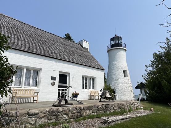 These photo shows a shorter, more conical white lighthouse with a black lantern room at the top. This lighthouse is built near a rocky shoreline, as evidenced by the stone wall or foundation visible in the foreground. The associated building is a white house with a distinctive slate or shingle roof, and there are wooden benches positioned around the property. An old anchor is displayed on the stone wall, adding to the maritime heritage theme. The setting appears more coastal and rugged compared…