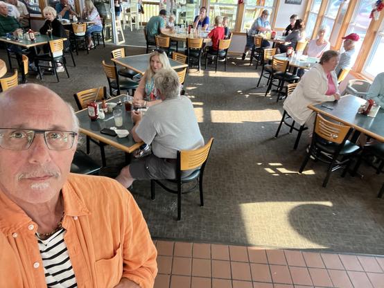 This photo shows the interior of a casual restaurant with a warm, welcoming atmosphere. In the foreground, a man [it’s me!] wearing glasses, an orange button-up shirt, and a black and white striped shirt underneath is taking a selfie. He has a gray beard and mustache.

The dining area features numerous wooden tables with matching wooden chairs that have horizontal slats in their backs. The floor appears to be tiled, and there’s carpeting in the seating area. Large windows along one side let in …