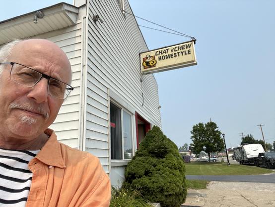 This photo shows a man [it's me!!] taking a selfie in front of a small, local restaurant. He's wearing glasses, has a gray goatee, and is dressed in an orange/coral colored shirt or jacket over what appears to be a striped shirt underneath.

Behind him is a white building with horizontal siding that houses "Chat 'N Chew Homestyle" restaurant, as indicated by the hanging sign with a coffee cup logo. The building is a converted residential structure or small commercial building typical of family-…