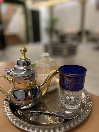 The photo shows a beautifully arranged tea service on a round silver tray.
	•	On the left is a small, ornate teapot made of polished silver with gold detailing on the handle, spout, and lid finial.
	•	Next to it is a clear glass cup decorated with an intricate cobalt blue and gold band around the top.
	•	Behind the glass is a white ceramic sugar container with golden crackle-like patterns, topped with a small glass knob.
	•	A silver spoon rests neatly on the tray in front of the glass.

The bac…