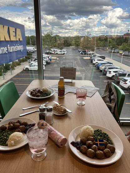 This photo shows a meal setup at an IKEA cafeteria. Three plates of food are on the table, each featuring Swedish meatballs with brown gravy, mashed potatoes, green peas, and lingonberry sauce. One plate has a small Swedish flag as a garnish. There are also glasses of pink sparkling drink with ice, a bottle of apple juice, wrapped cookies, and a slice of layered dessert on a small plate. Salt and pepper shakers are on the table as well.

The table is positioned near a large window overlooking t…