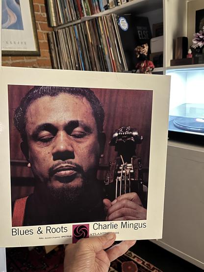 This photo shows someone holding a vinyl record cover of Charlie Mingus – Blues & Roots, a classic jazz album released on Atlantic Records. The album cover features a close-up portrait of Charles Mingus with his eyes closed, holding the neck of his upright bass.

In the background, you can see part of a cozy room with a record collection on shelves, a turntable with a record spinning, and some decorative items including flowers and a figurine. A framed artwork is visible on the left wall, and t…