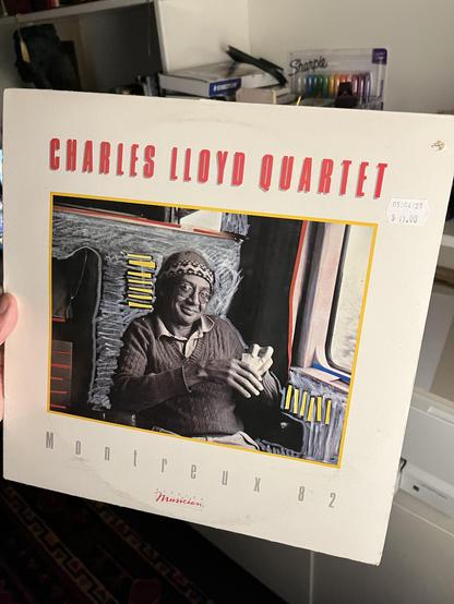 The photo shows someone [that would be me] holding a vinyl record album. The cover features the Charles Lloyd Quartet with the title “Montreux 82” written at the bottom.

The design has a mostly white background with bold red text at the top for the artist’s name.

In the center is a framed image of Charles Lloyd seated casually, smiling, wearing a knit cap and a sweater. The photo has some drawn or painted artistic embellishments around it, giving it a stylized look.

The bottom right corner o…