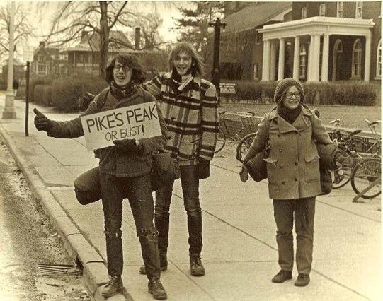 This is a black and white photograph from the 1970s showing three young people on a college campus. Two people on the left are hitchhiking, with one holding a sign that reads "PIKES PEAK OR BUST!" - a reference to the famous mountain in Colorado. The person [it me] holding the sign has their thumb out in the classic hitchhiking gesture. A third person stands to the right, and all three are dressed in typical 1970s casual wear including jackets and jeans.

In the background, you can see campus-s…