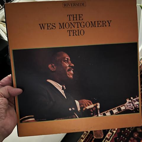 The photo shows someone [it me!] holding a vinyl record album titled “The Wes Montgomery Trio”, released on the Riverside label. The album cover is predominantly orange with a rectangular black-and-color photograph of Wes Montgomery, the legendary jazz guitarist, smiling while playing his guitar. He is dressed in a suit and appears to be performing. The design is clean and classic, with bold black text above the photo and the Riverside label logo at the top.