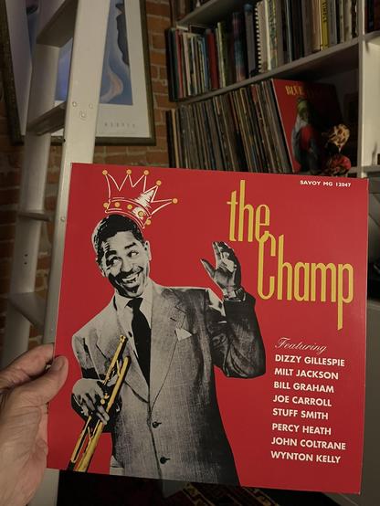 This photo shows someone [that would be me, Claude] holding a jazz album titled "The Champ" on the Savoy Records label (catalog number MG 12047). The album cover features a striking red background with a black and white photograph of a smiling musician [Dizzy Gillespie!] in a suit holding a trumpet, with a golden crown illustration above his head - fitting the "Champ" theme.

The album features an impressive lineup of jazz legends, as listed on the cover: Dizzy Gillespie, Milt Jackson, Bill Gra…