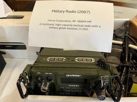 This photo shows a military tactical radio system on display. The main item is a Harris Corporation RF-7800M-MP multiband radio in military olive drab green, with “FALCON III” visible on the front panel. According to the information placard, this is from 2007 and comes with a military-grade H-250 handset.
The radio features various connectors and ports on the front, including what appears to be GPS and USB/KDU connections. There’s a coiled handset cord and a traditional military handset visible…